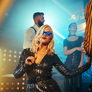Nightclub scene with woman in sparkly black outfit and sunglasses in foreground. Blue lighting and blurred figures in background.