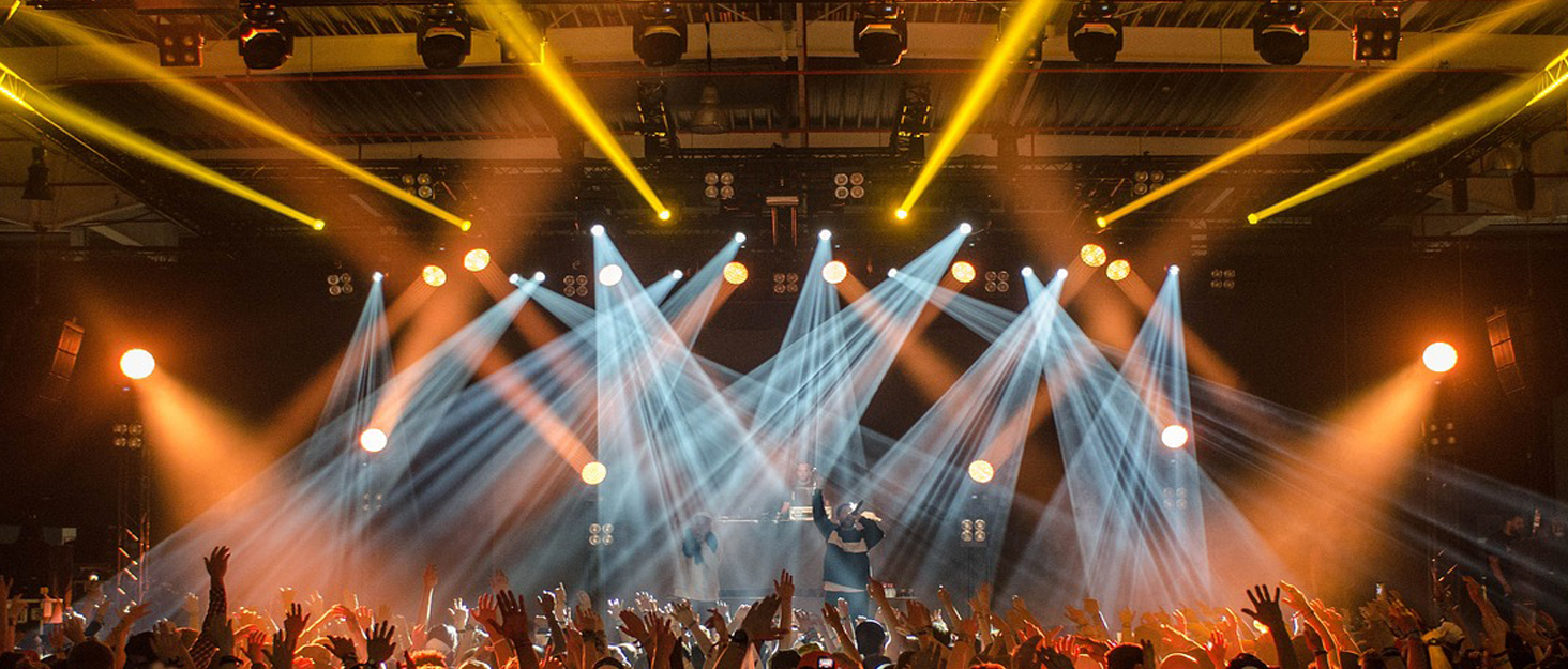 Concert stage with dramatic lighting. Multiple spotlights create crisscrossing beams in yellow and white. A silhouetted performer stands center stage with arms raised as a crowd cheers.