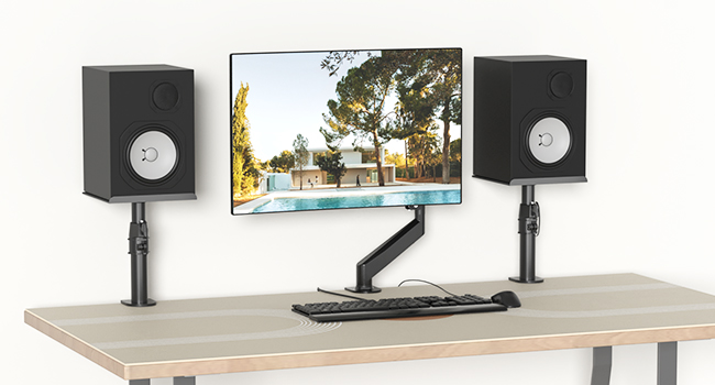 Desktop setup with dual speakers on stands flanking a monitor on adjustable arm mount, arranged on light wooden desk surface.