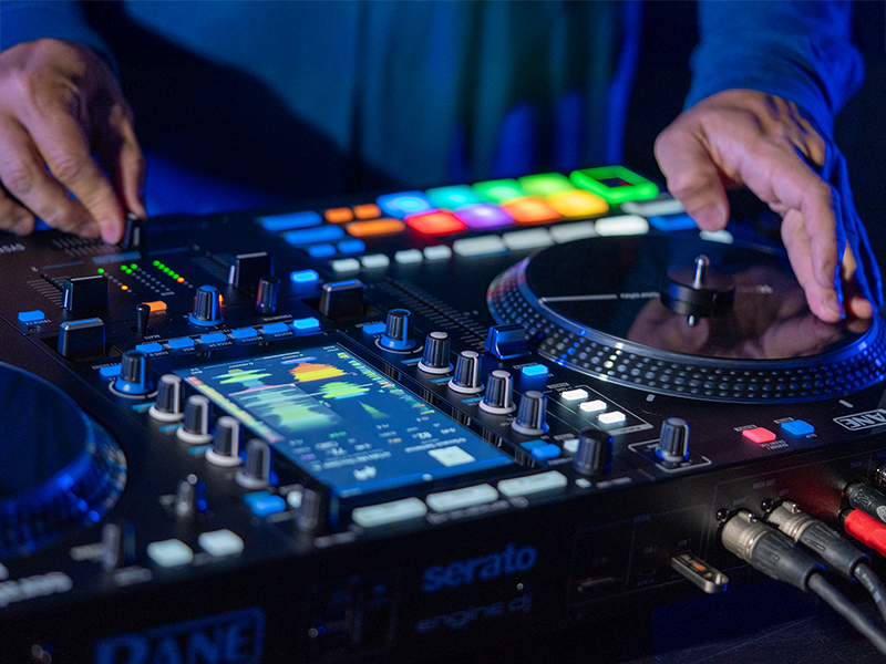 Professional DJ controller with illuminated multicolor performance pads, mixer controls, and turntable deck in dark setting with hands visible operating equipment.