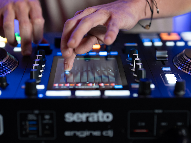 Close-up view of DJ equipment with mixing controls, turntables, and interface showing 'Serato' software. Various knobs, faders and digital displays visible in blue lighting.