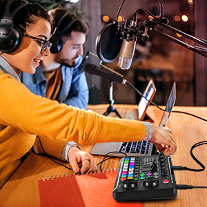 Two people in a recording studio, using laptops and audio equipment. Woman in yellow sweater operates mixing board, both wear headphones near microphone.