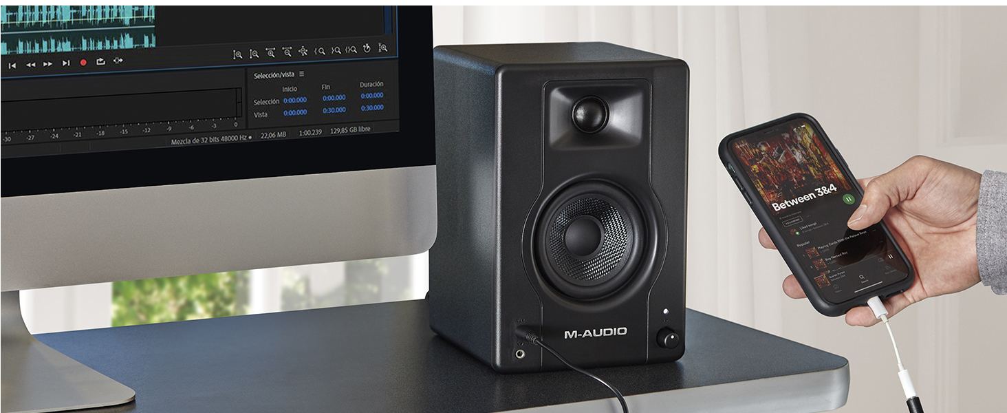 Black studio monitor speaker on desk next to computer monitor, with hand holding smartphone displaying control interface.