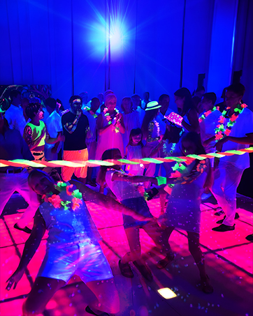 Crowded nighttime event or venue with purple and pink neon lighting. Large crowd of people gathered under illuminated structures. High-energy entertainment environment with vibrant colored stage lighting.